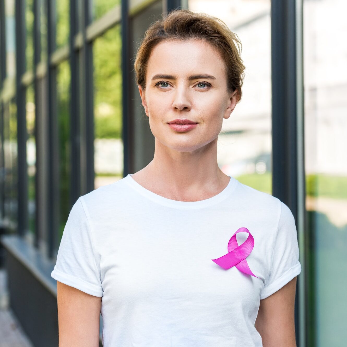 pensive middle aged woman with pink ribbon standing on street, breast cancer awareness concept
